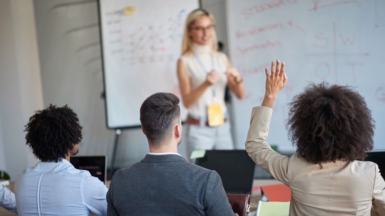 Claim Your Seat - 8 Reasons To Speak Up In The Workplace - a group of people in a work presentation with one person raising their hand.